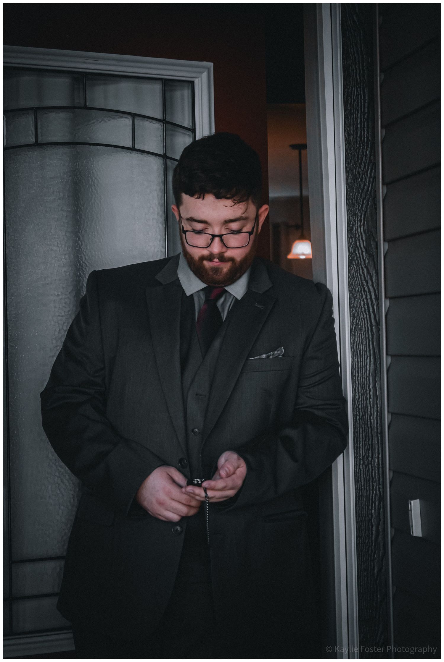 A bearded man in a dark suit adjusts his cufflinks while standing in a doorway, dressed for a formal occasion.