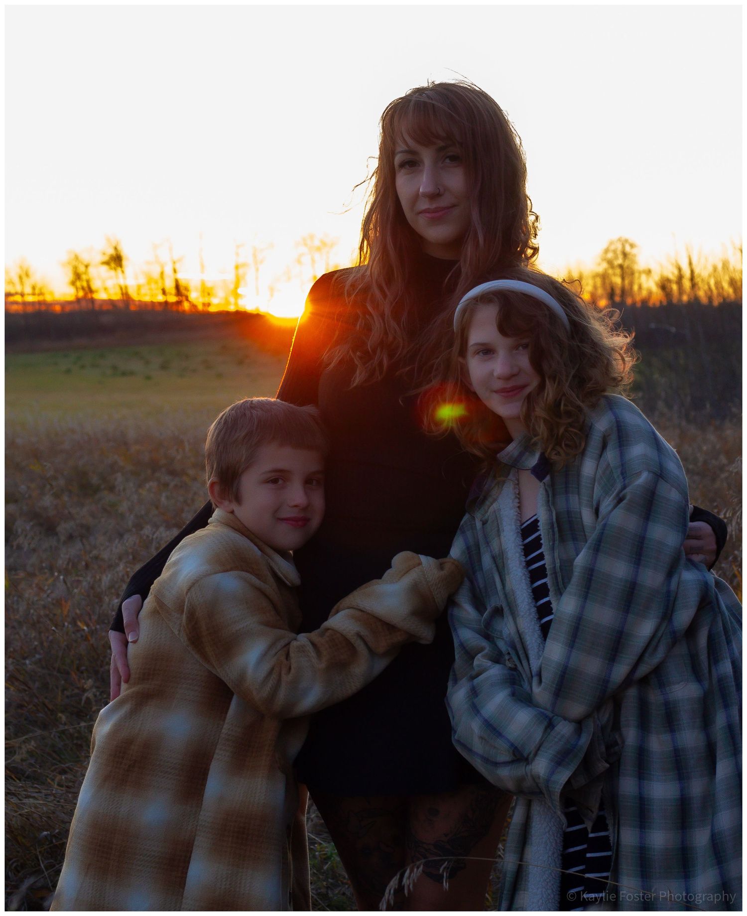 A woman and two children pose together in a field at sunset with golden light glowing behind them.