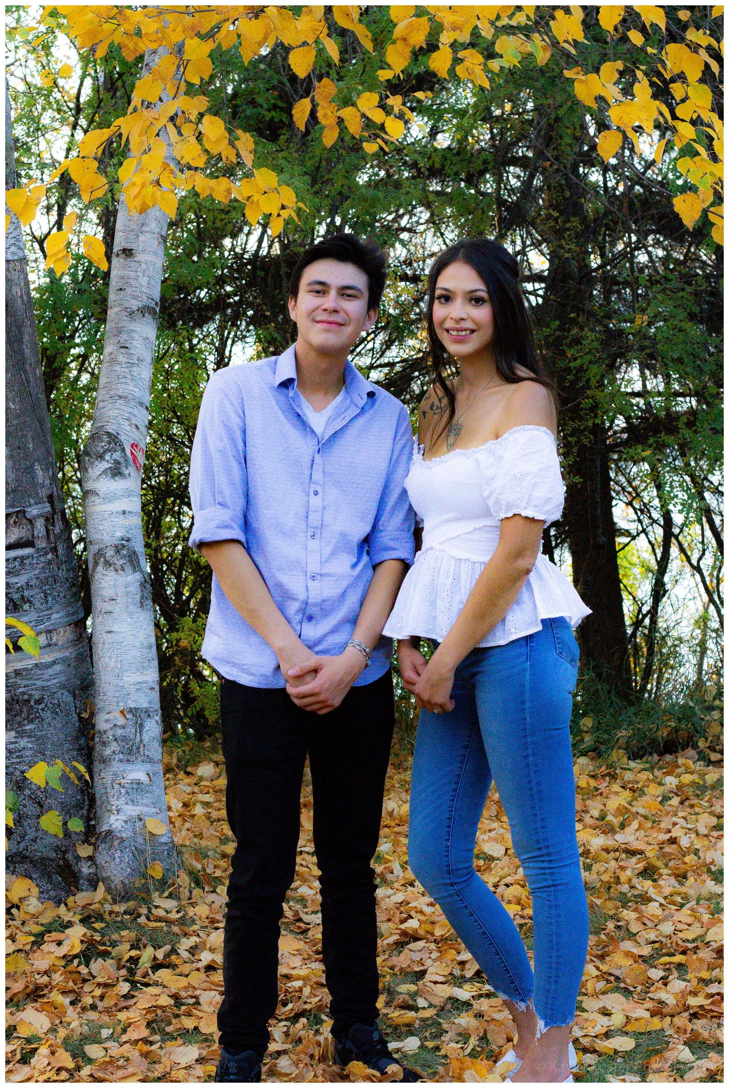 Young couple standing among fall foliage near birch trees, man in blue shirt and woman in white top with jeans.