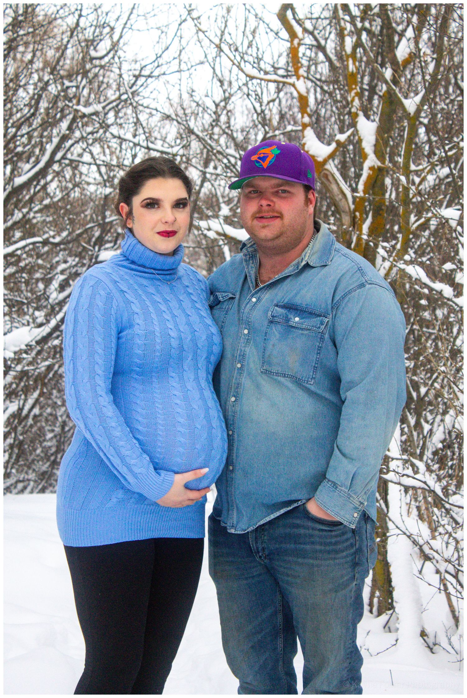 Pregnant couple posing outdoors in winter snow, woman in blue turtleneck sweater cradling baby bump.