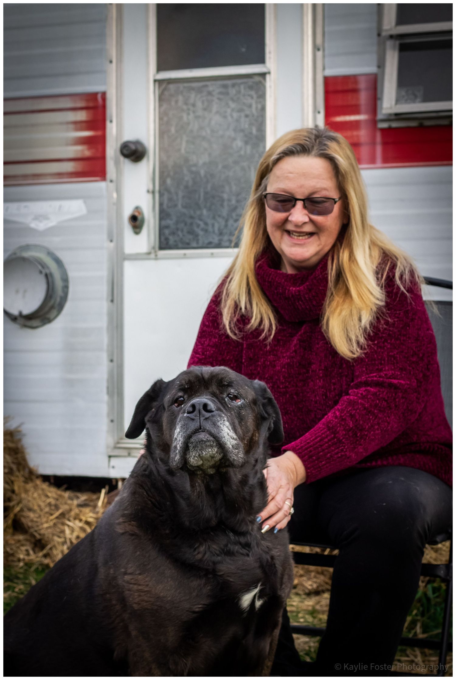 Woman in red sweater smiling with large black dog in front of vintage trailer outdoors.