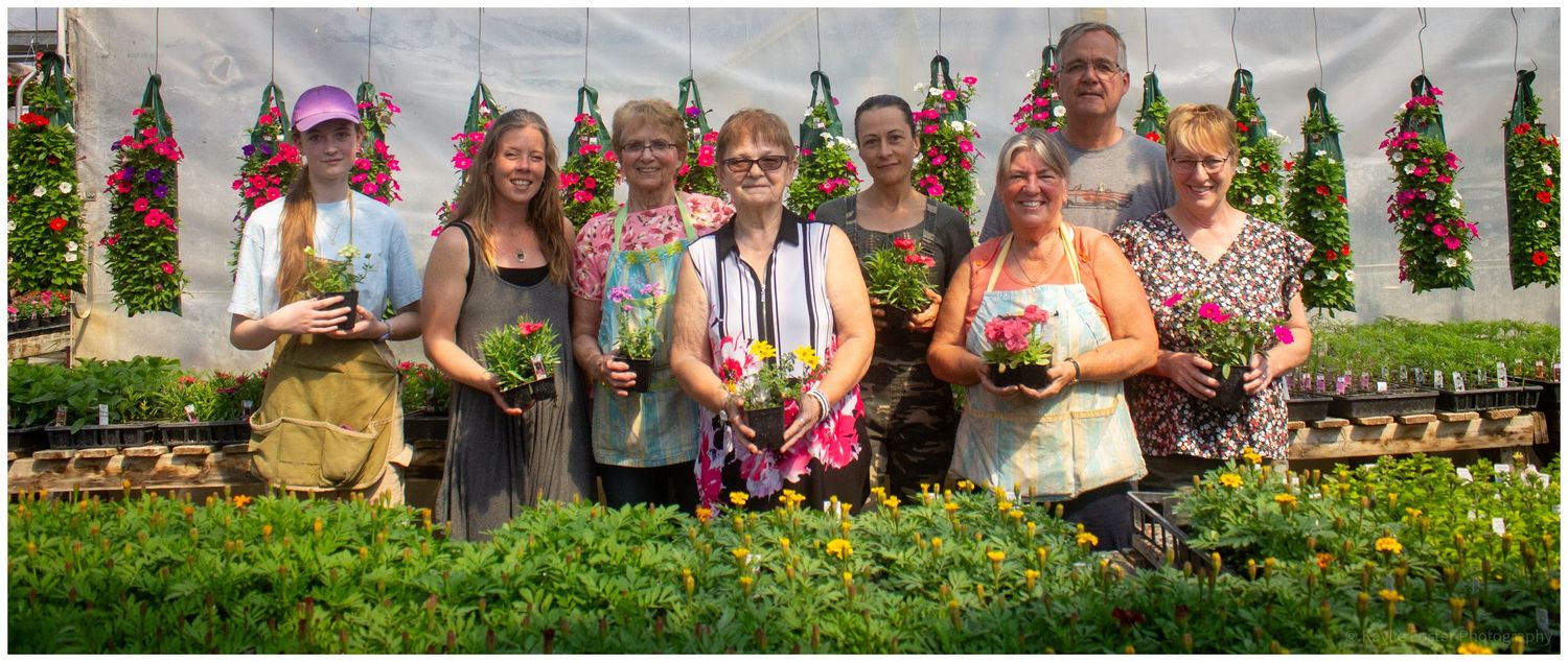 Group of people holding colorful flowers and plants inside a greenhouse nursery filled with hanging baskets.