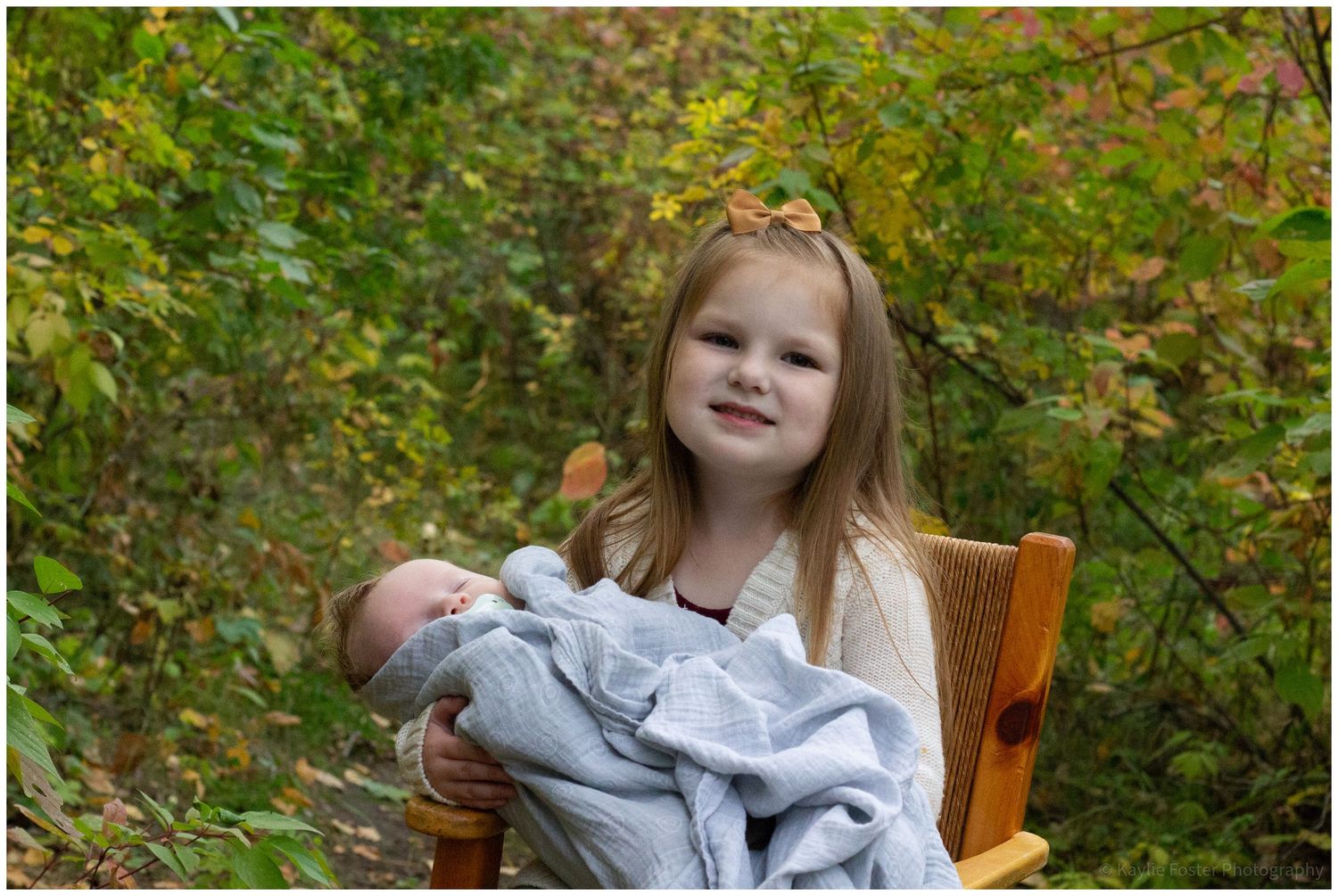 Young girl sitting in wooden chair holding a newborn baby wrapped in a blue blanket outdoors.
