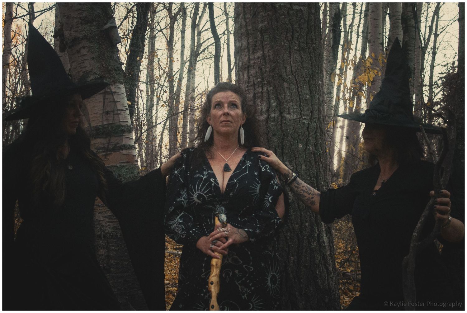 Three women dressed as witches in dark gowns standing among birch trees in an autumn forest.