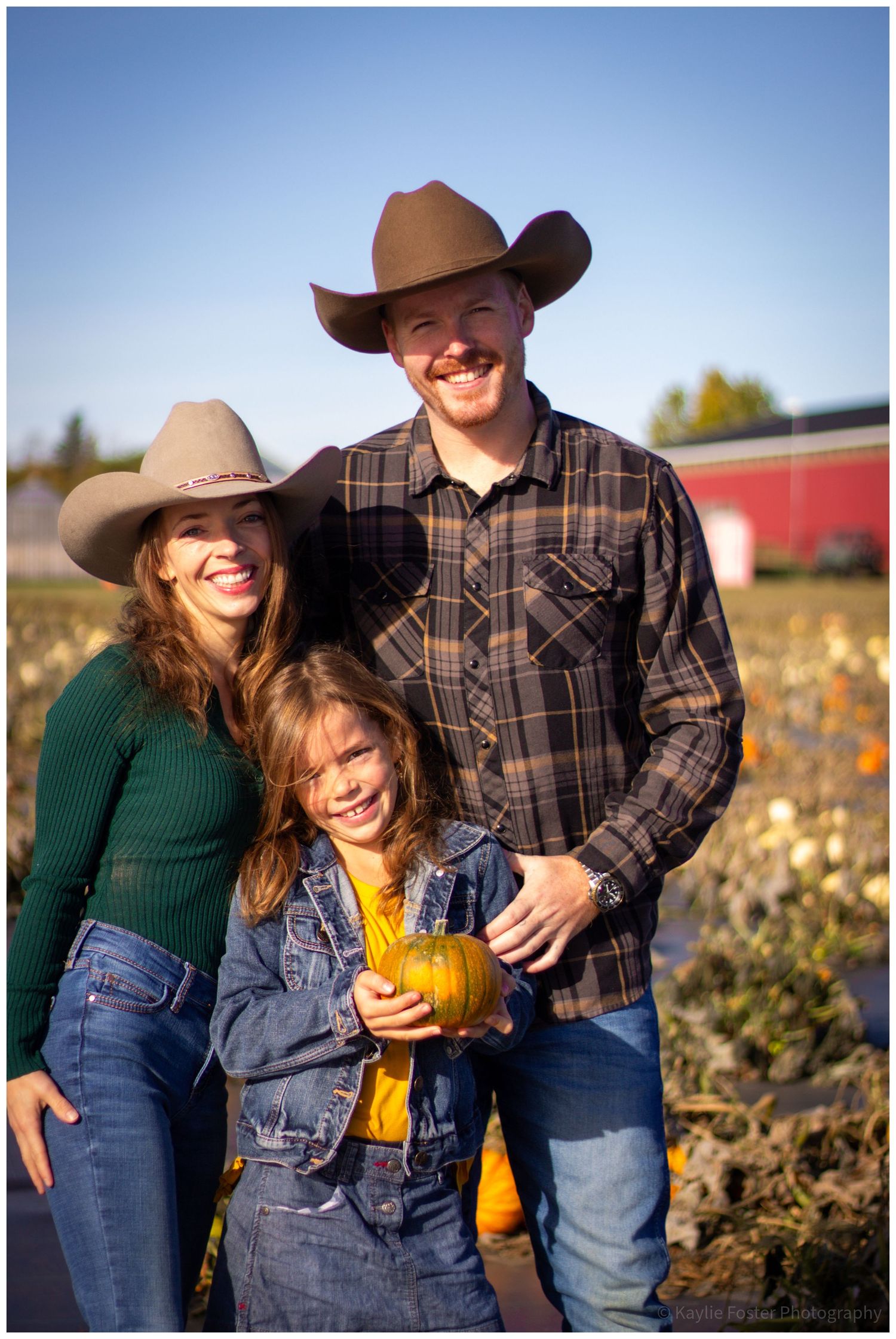 Family of three in cowboy hats smiling at a pumpkin patch, child holding small pumpkin, sunny autumn day.