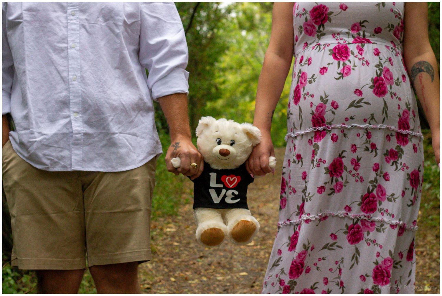 Couple holding a teddy bear with a LOVE shirt during a maternity photo shoot outdoors.