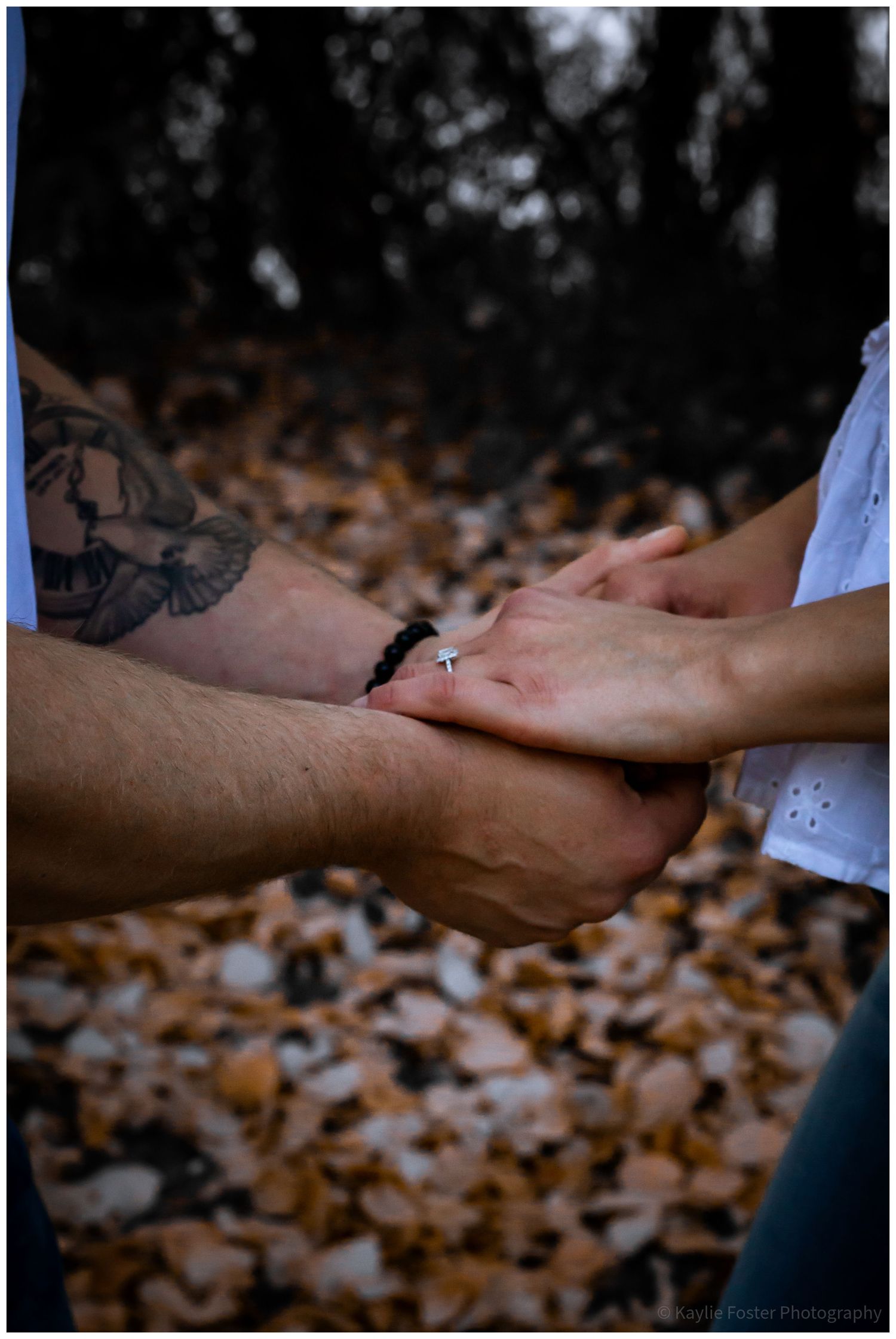 Couple holding hands outdoors in autumn leaves, man with tattoos and bracelet, woman wearing a cross ring.