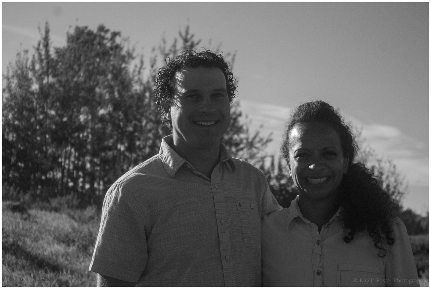 Black and white photo of a smiling man and woman posing outdoors with trees in the background.