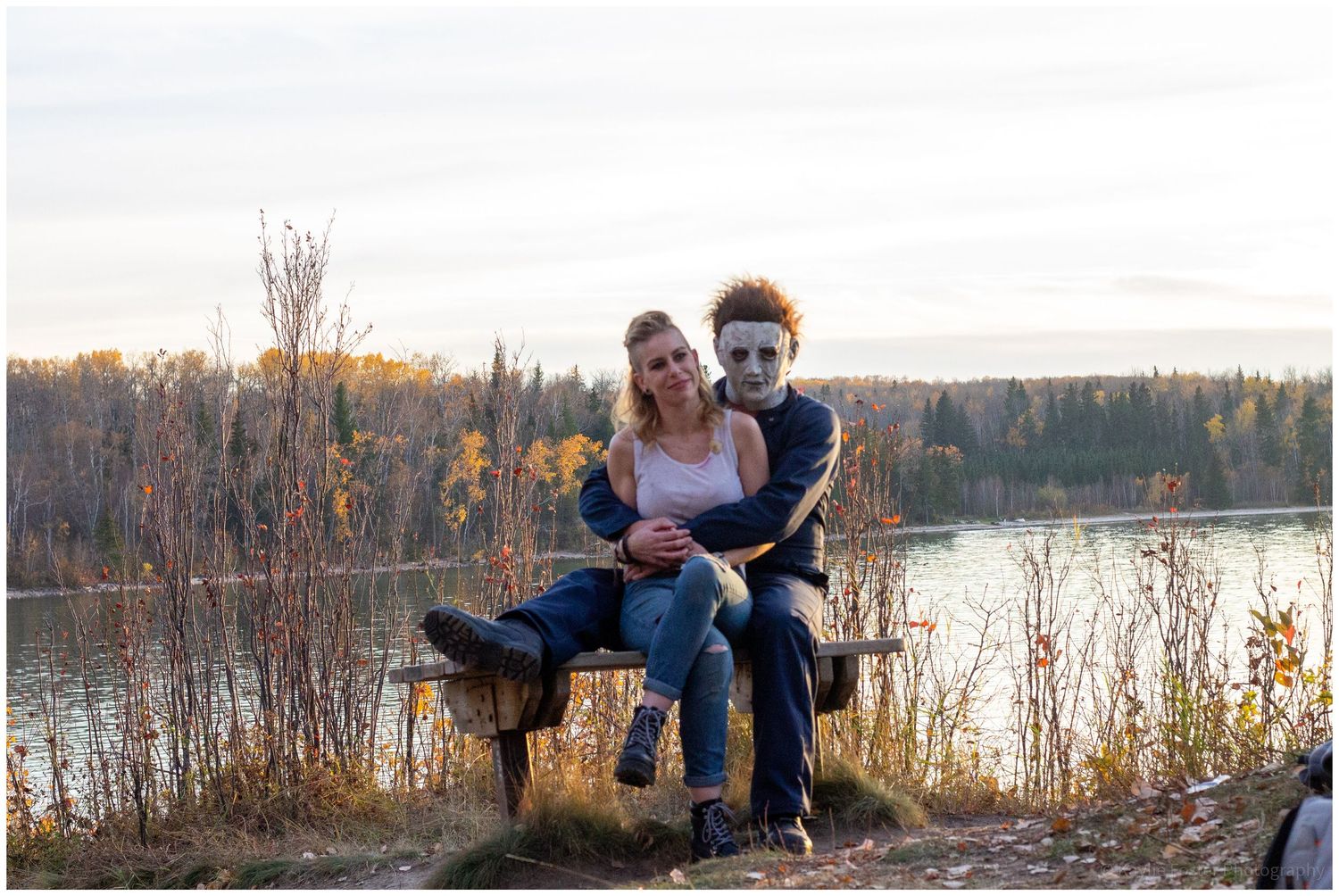 Couple posing on a bench by a lake in autumn, one wearing a Halloween Michael Myers mask, surrounded by fall foliage.