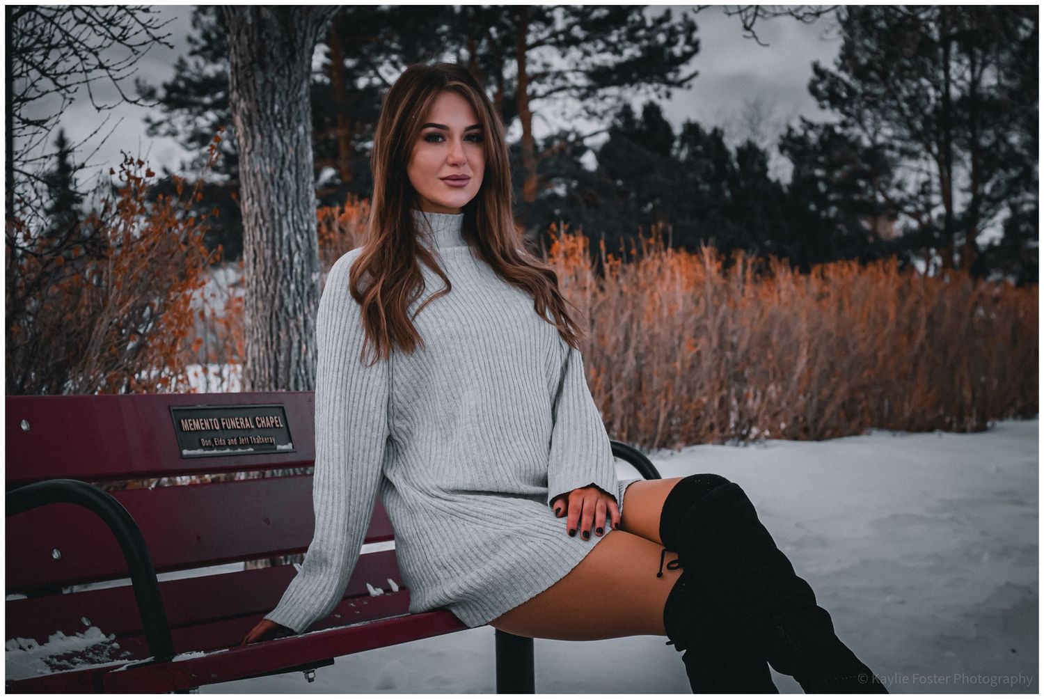 Attractive young woman in gray sweater dress and knee-high boots sitting on snowy park bench.