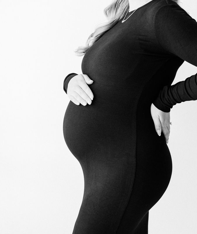 Black and white maternity photo of pregnant woman in black bodysuit cradling baby bump against white background.