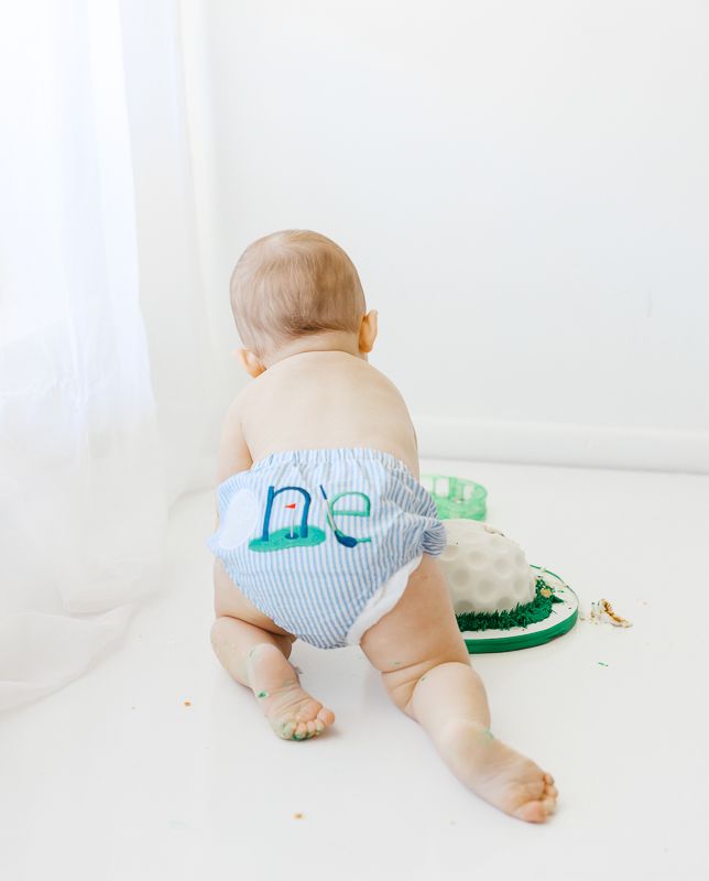 Baby in blue seersucker diaper cover with golf embroidery crawling toward a smash cake on white background.