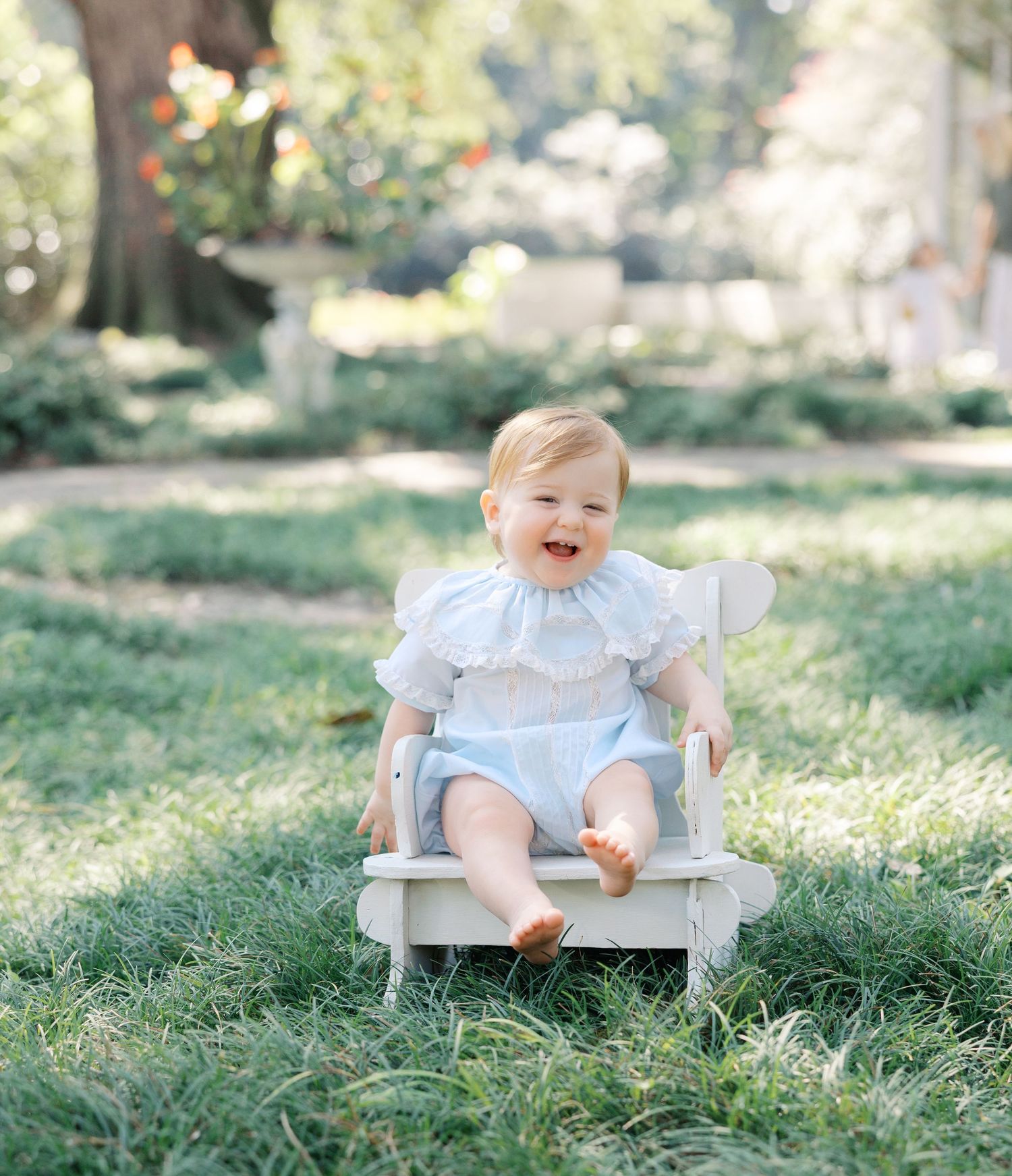 Happy baby in blue smocked outfit sitting in white Adirondack chair on green grass.