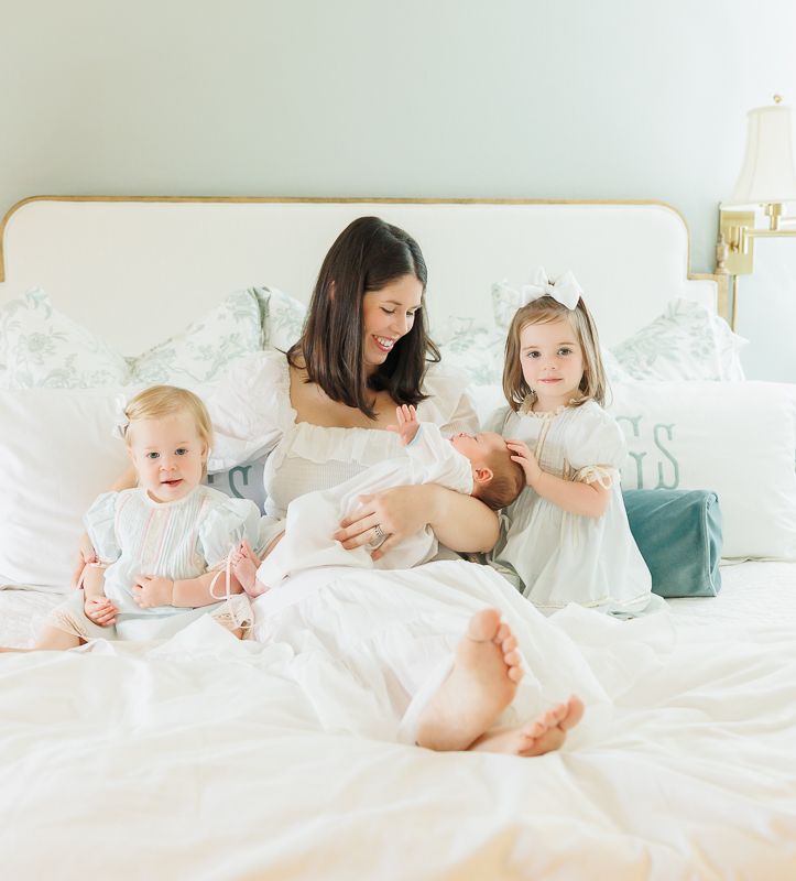 A mother sits on a white bed holding a newborn, flanked by two young daughters in white dresses.
