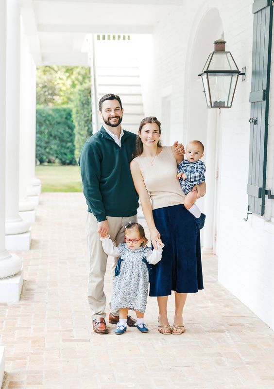 A smiling family of four poses on a white columned porch, parents holding two young children in preppy attire.