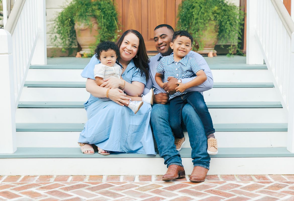 Happy African American family of four sitting on front porch steps, parents holding young children, wearing coordinated blue outfits.