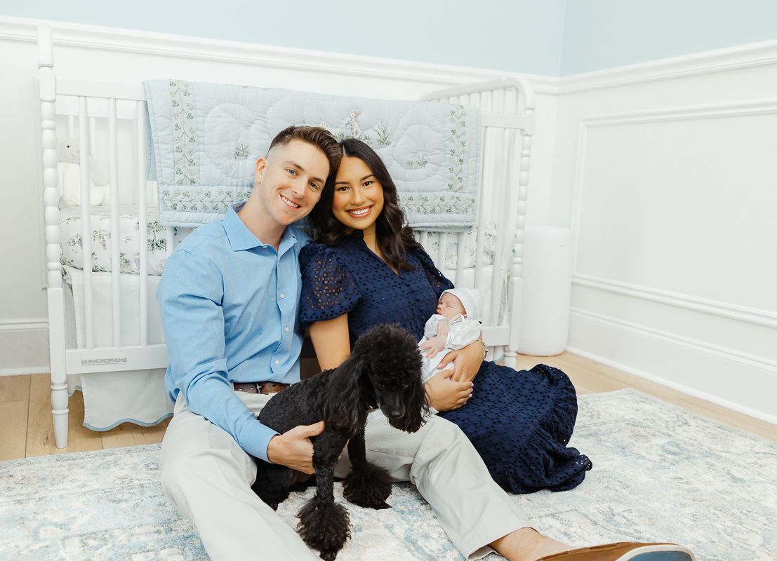 Happy couple sitting on floor with newborn baby and black dog in elegant nursery room.