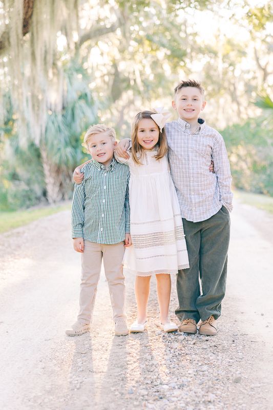 Three smiling children pose together on a sunlit path surrounded by moss-draped trees in coordinated pastel outfits.
