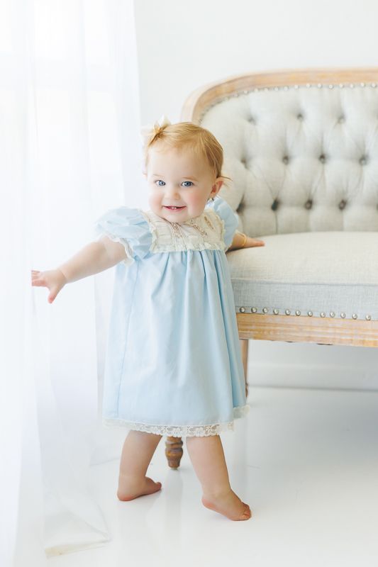 Smiling baby girl in light blue lace dress standing by elegant tufted sofa in bright white room.