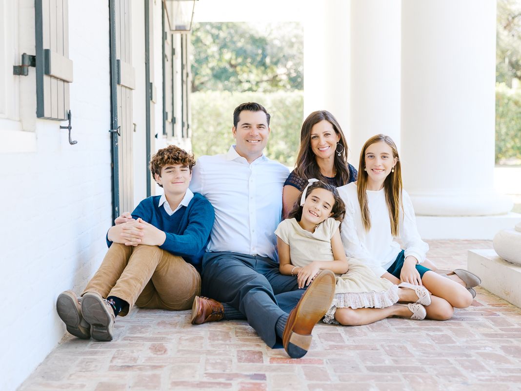 A happy family of five poses together on brick steps outside a white building for a portrait photo.
