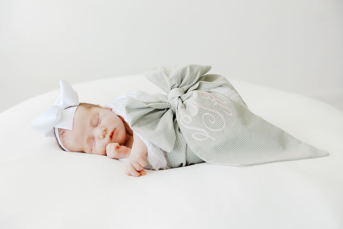 Newborn baby swaddled in a gray monogrammed blanket with a white bow headband, sleeping peacefully on white background.