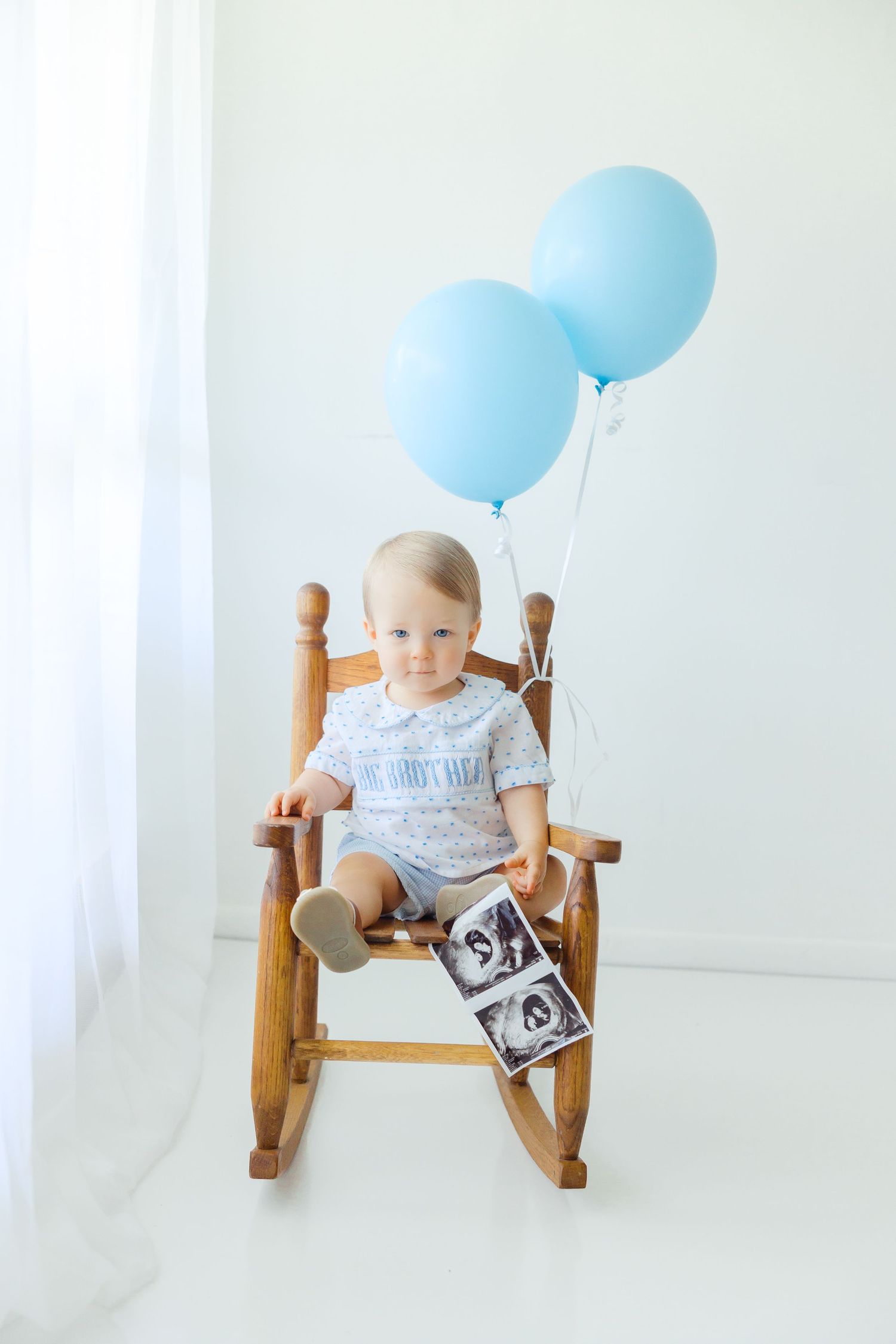 Adorable baby sitting in wooden rocking chair holding ultrasound photos with blue balloons tied above.