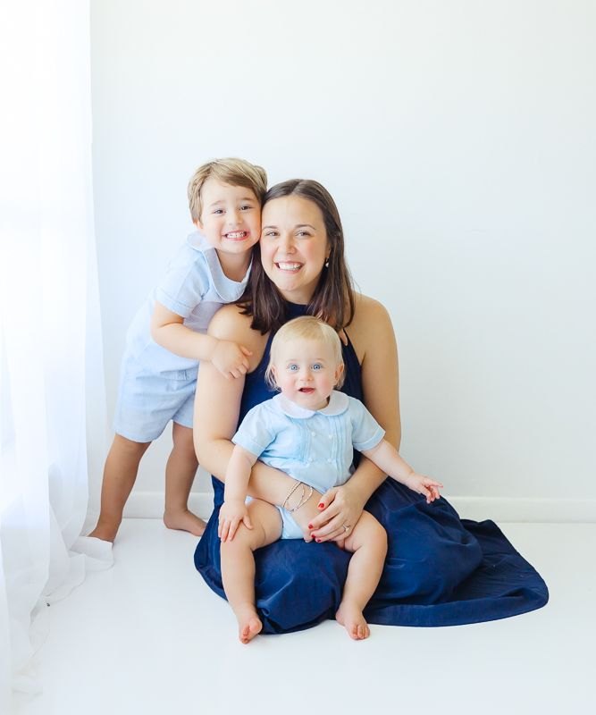 A smiling mother sits on a white floor with two young boys in blue outfits, posing for a bright family portrait.