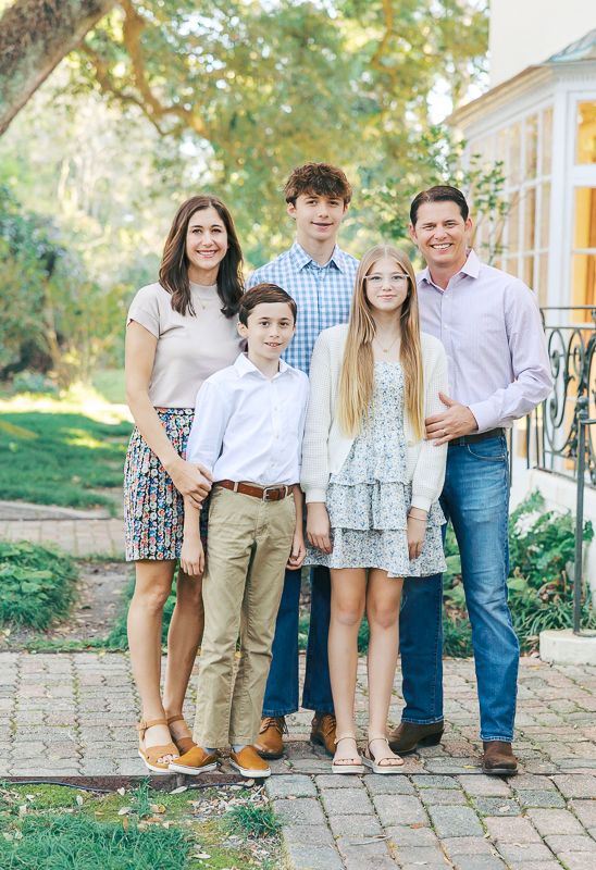 A happy family of five poses outdoors on a brick path, dressed in smart casual attire, surrounded by lush greenery.