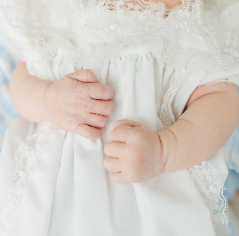 Close-up of a baby in a white lace christening gown, with tiny chubby hands gently clasped together.