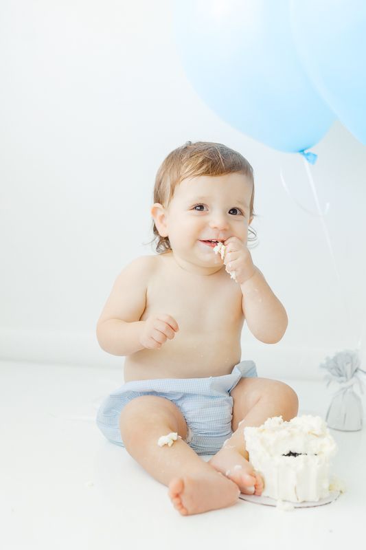Baby boy in blue diaper smashes first birthday cake, smiling with frosting on face, blue balloon in background.