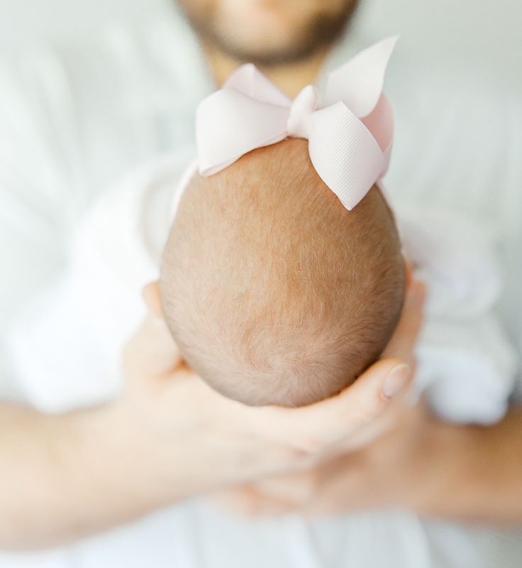 A father gently holds his newborn baby girl, showing the back of her head adorned with a pink bow.