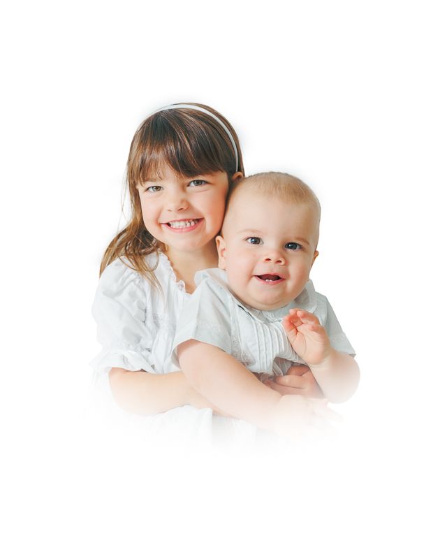 A smiling young girl in white dress hugging a happy baby, both posing for a bright portrait photo.