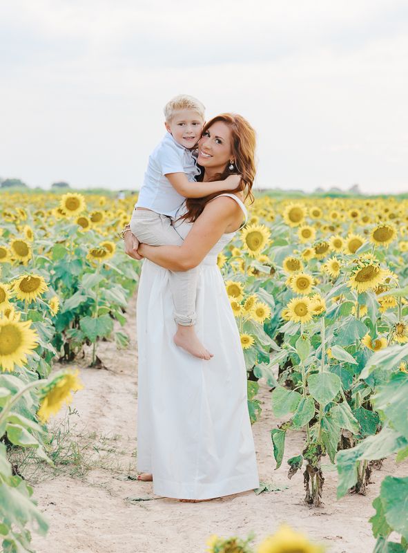 Woman in white dress holding a toddler boy in a sunny sunflower field, smiling warmly for a portrait photo.