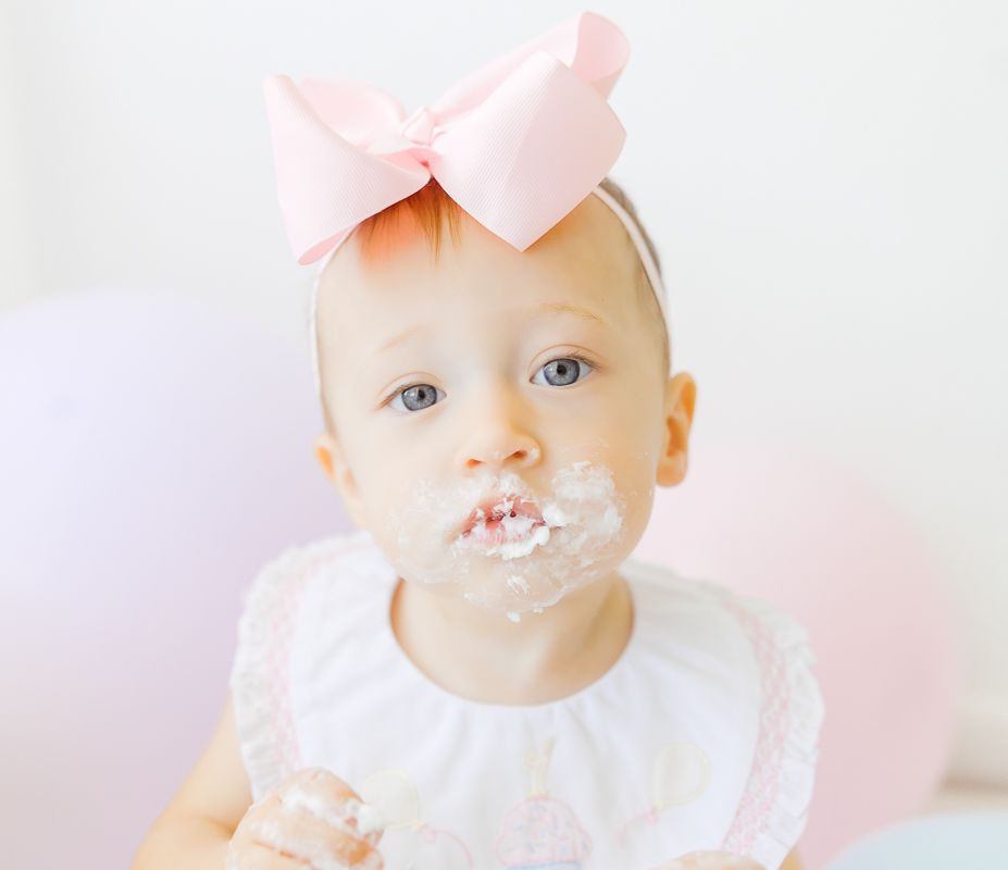Baby girl with pink bow headband eating birthday cake with frosting covering her mouth.