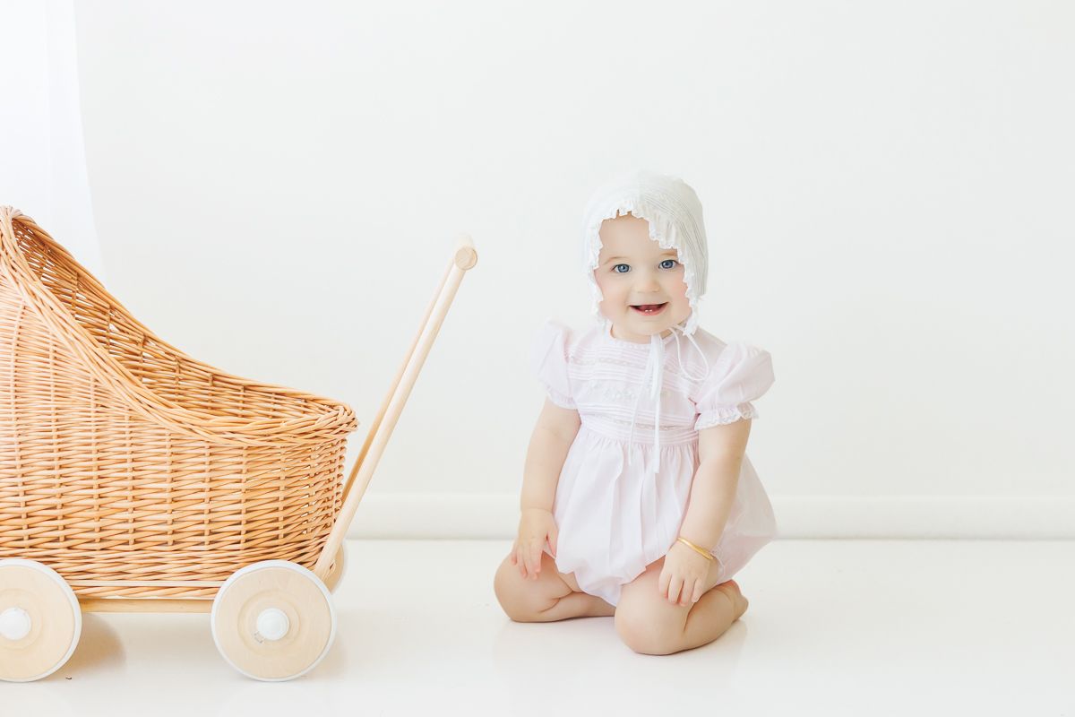 Smiling baby girl in pink romper and white bonnet sitting on floor next to a wicker doll pram.