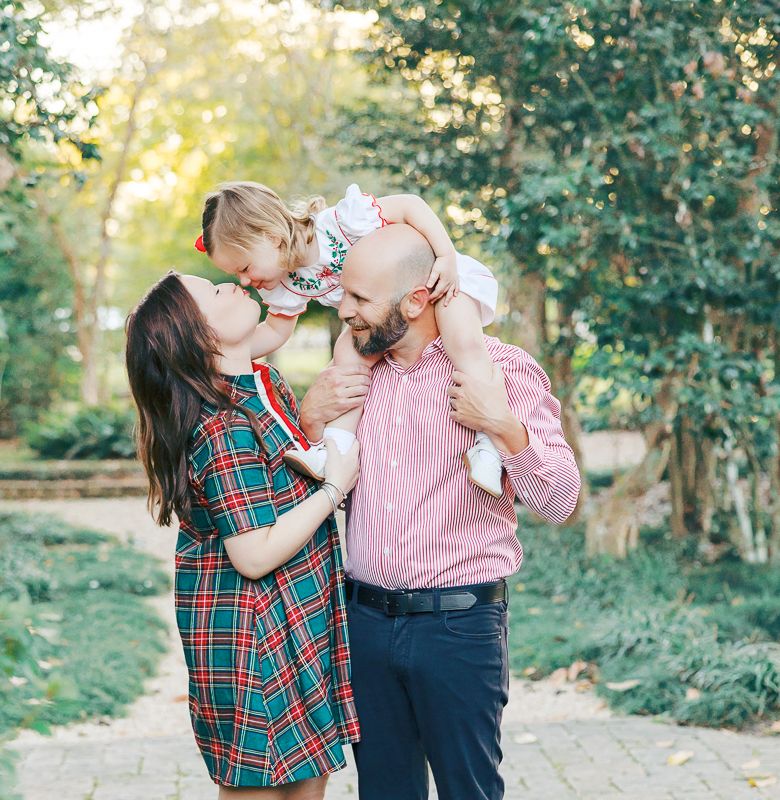 A happy family with a bald father, brunette mother in plaid dress, and toddler girl sharing a kiss outdoors.