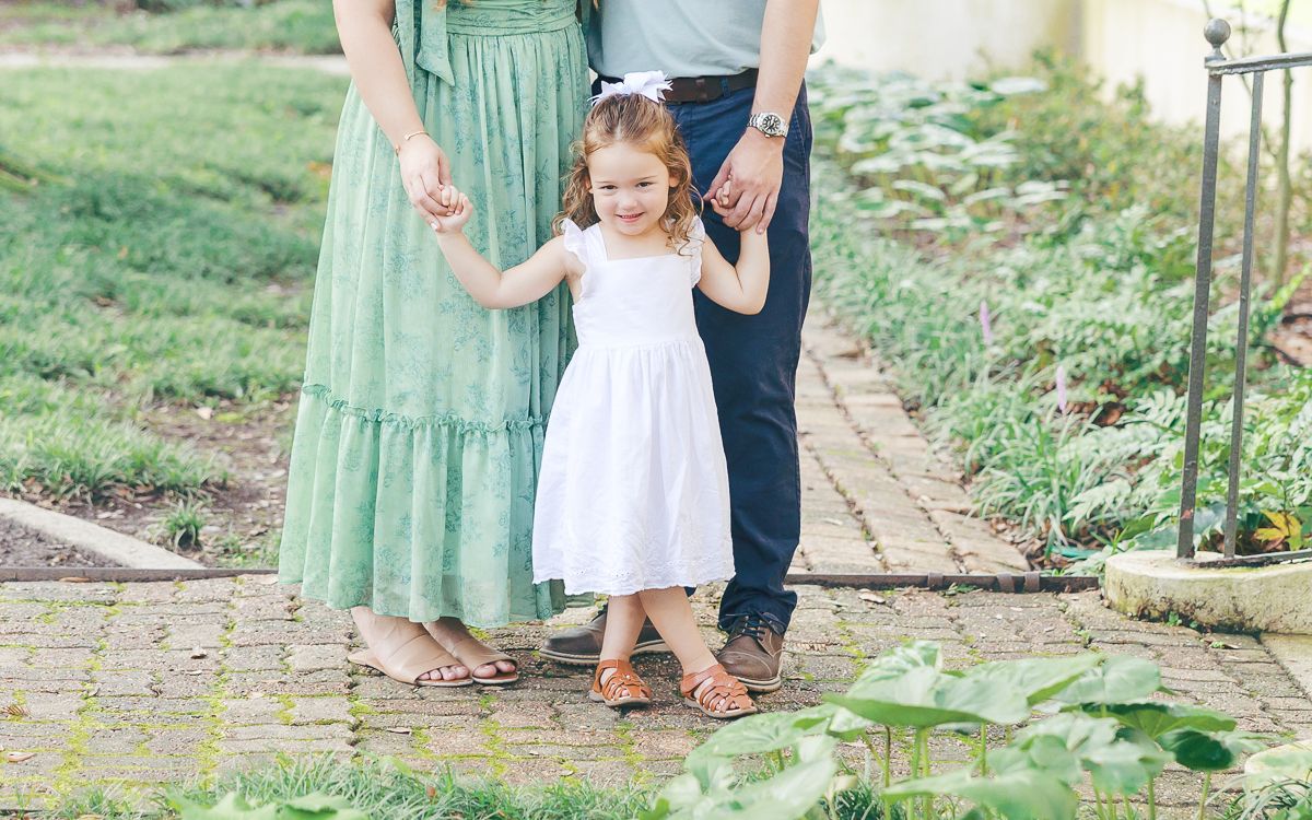A young girl in a white dress holds hands with her parents in a garden setting.