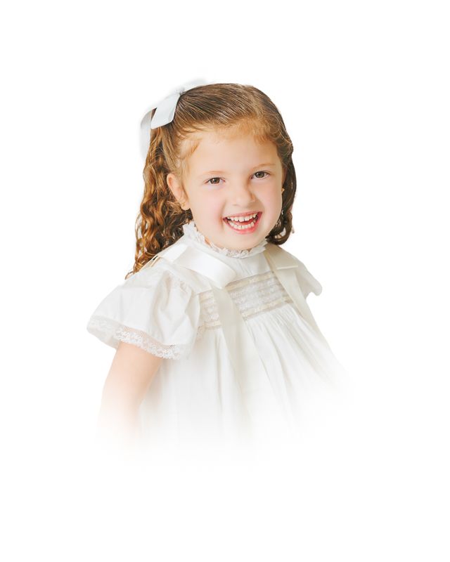 Smiling young girl with curly hair wearing a white dress and bow, posed against a white background.