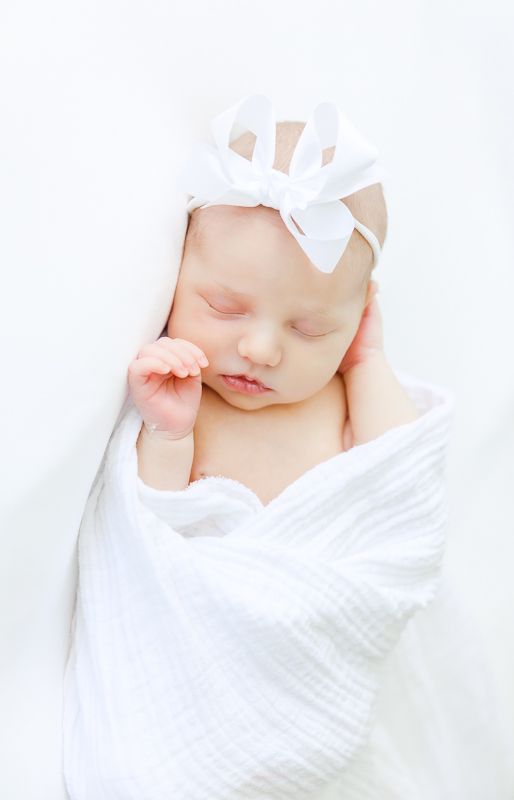 Peaceful newborn baby sleeping swaddled in white muslin wrap with white bow headband against white background.