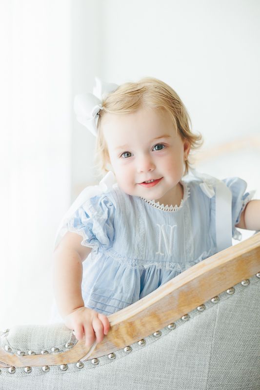 Smiling toddler girl in blue dress with pearl necklace and white bow stands beside a gray upholstered chair.