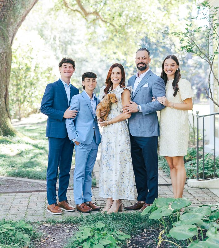 Family of five posing outdoors in elegant attire with a golden dog in a lush garden setting.