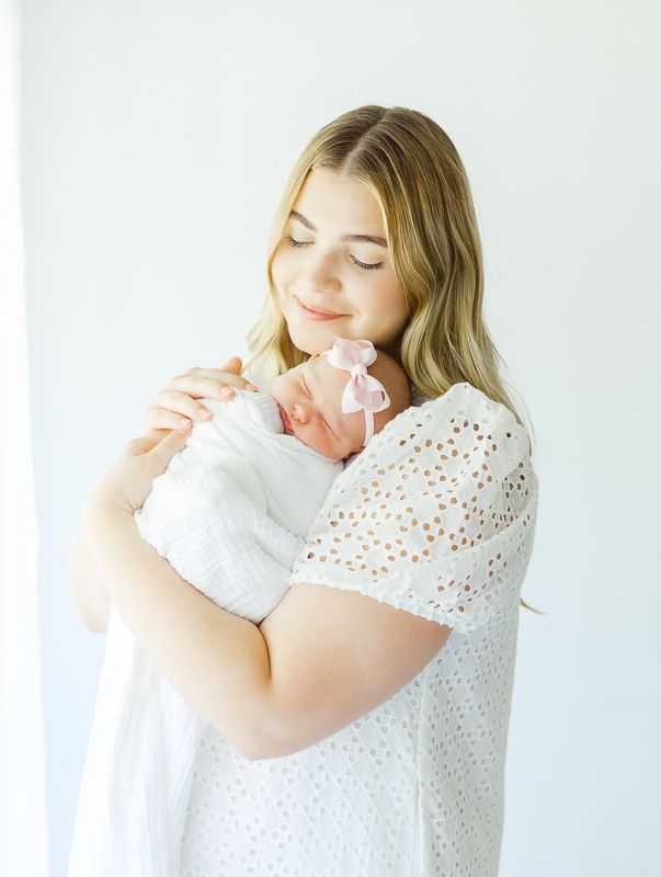 A young mother in a white lace dress tenderly holds her swaddled newborn baby with a pink bow in a bright studio.