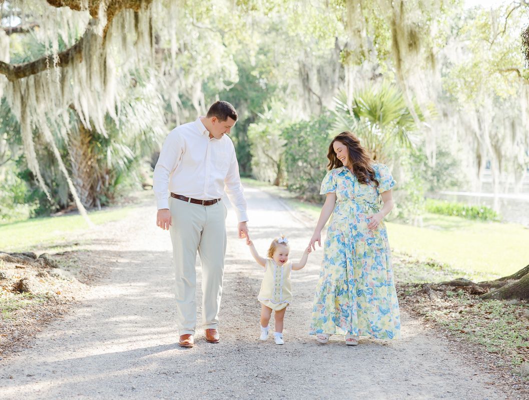 Family of three walks hand-in-hand on a sunlit path beneath moss-draped oak trees in a lush Southern garden.