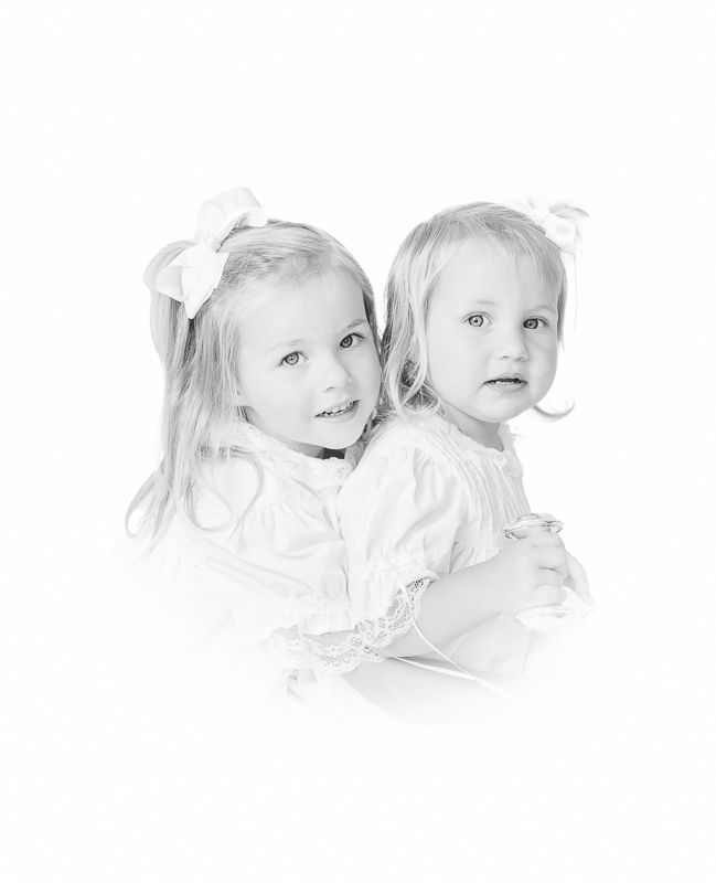 Black and white portrait of two young girls in white dresses with hair bows, posing together sweetly.