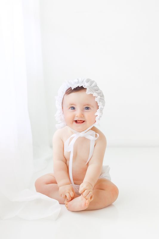 Smiling baby girl sitting up wearing a white bonnet and diaper in a bright white studio setting.