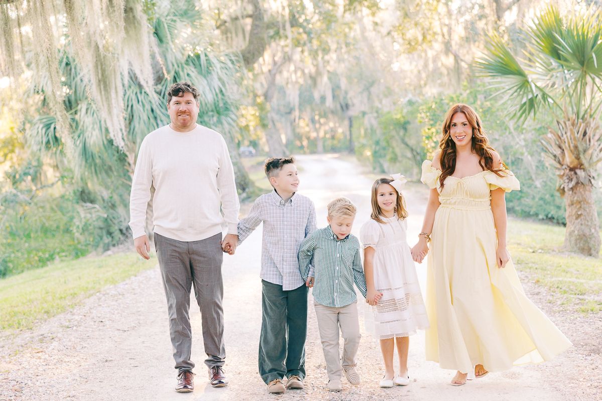 Family of five walking hand-in-hand on a sunlit path surrounded by lush trees in pastel outfits.