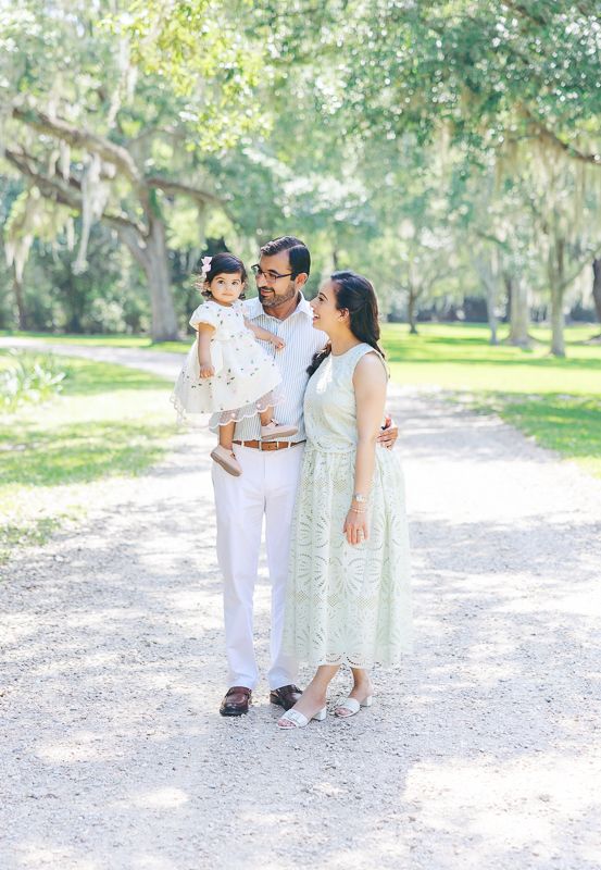Family of three poses on a sunlit gravel path surrounded by moss-draped trees, dressed in elegant white and mint outfits.