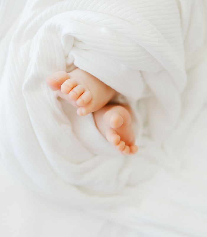 Newborn baby feet peeking out of a soft white swaddle blanket, close-up photography.