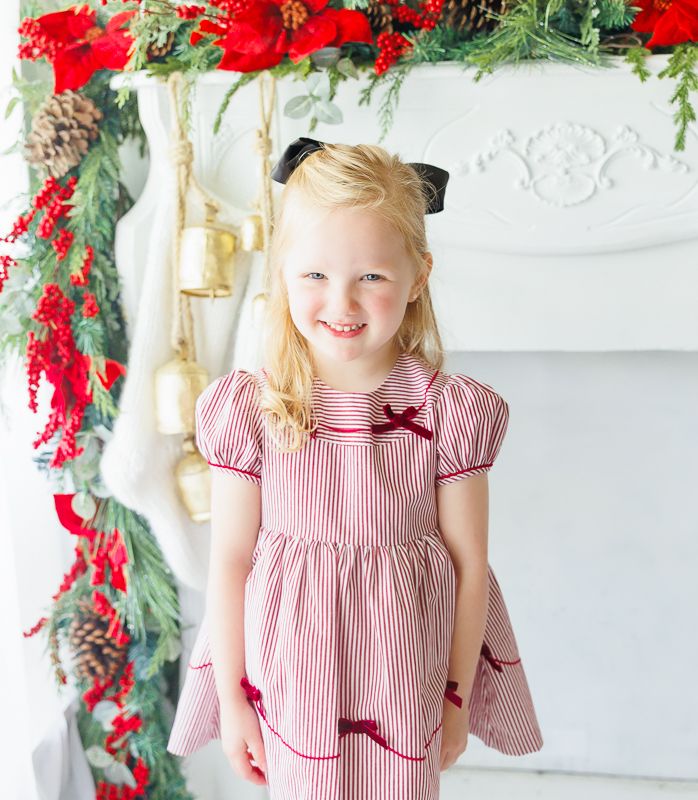 Young girl in red striped Christmas dress with velvet bows poses by festive garland-decorated fireplace mantel.