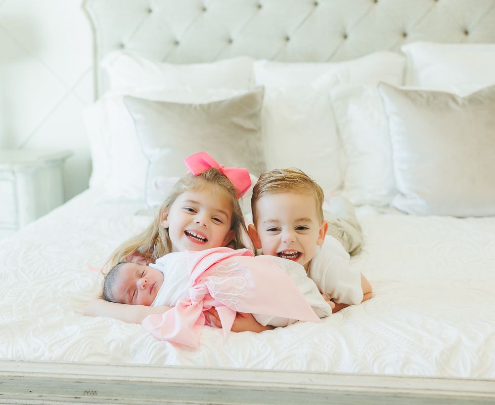Two happy siblings smile while holding their newborn baby sibling on a white bed with pillows.